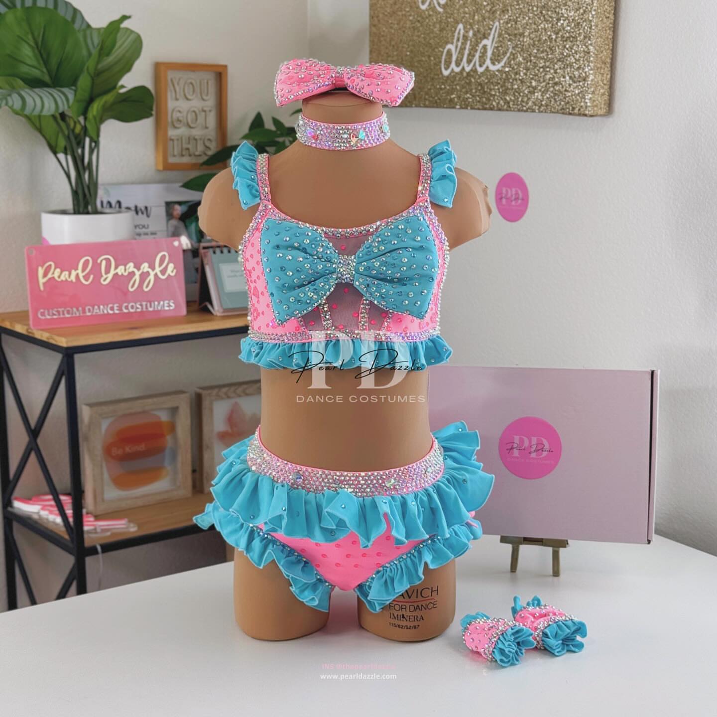 Turquoise and pink two-piece rhinestone dance costume with oversized bow top, crystal choker, layered chiffon ruffle bottoms, and matching hair bow displayed on mannequin.