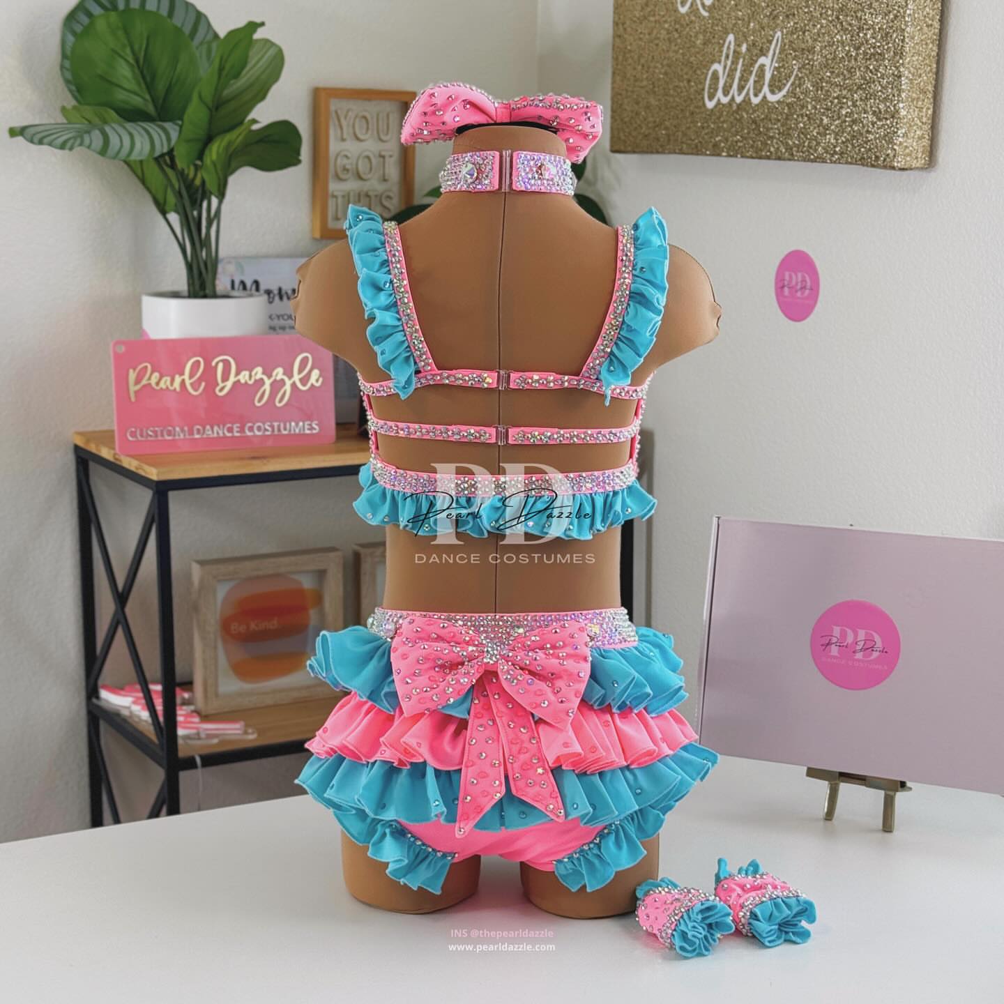 Turquoise and pink two-piece rhinestone dance costume with oversized bow top, crystal choker, layered chiffon ruffle bottoms, and matching hair bow displayed on mannequin.