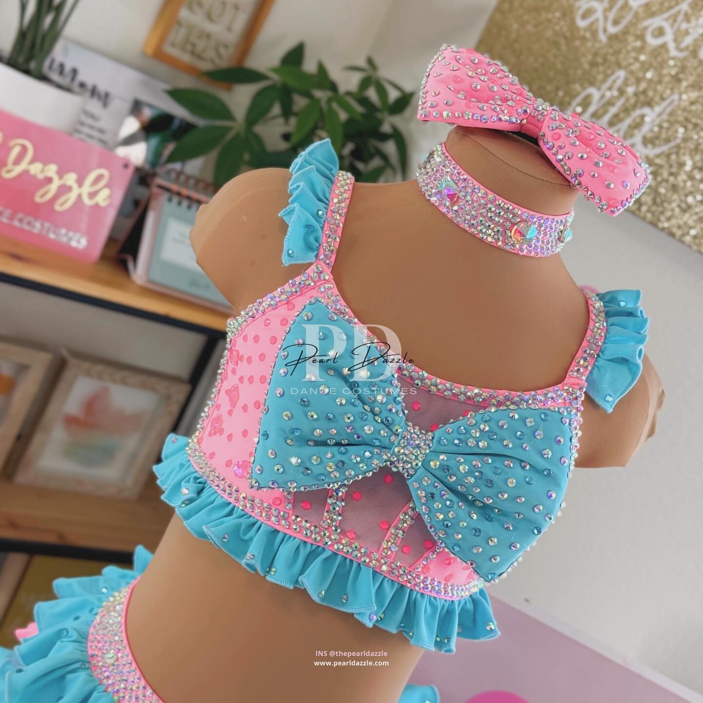 Turquoise and pink two-piece rhinestone dance costume with oversized bow top, crystal choker, layered chiffon ruffle bottoms, and matching hair bow displayed on mannequin.