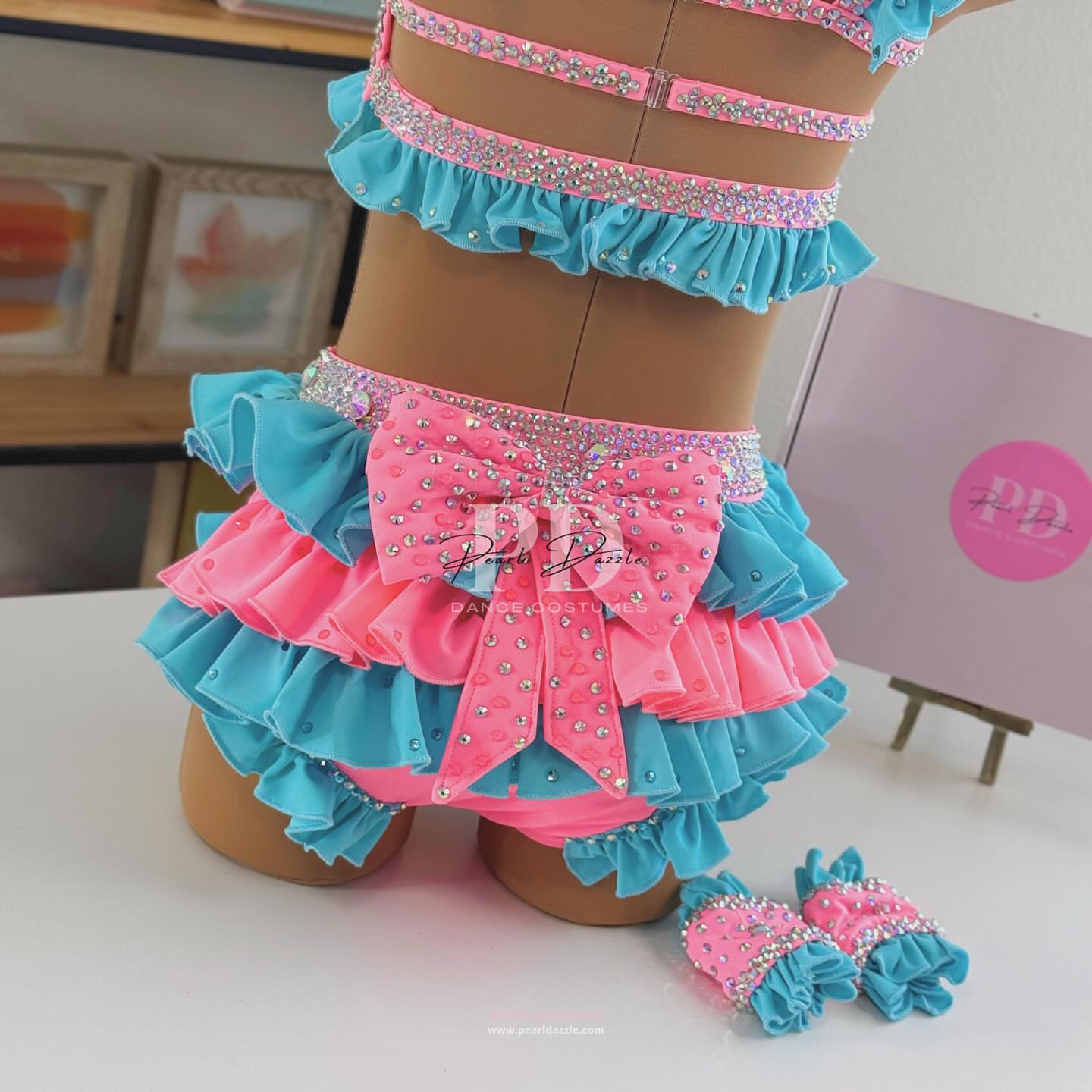 Turquoise and pink two-piece rhinestone dance costume with oversized bow top, crystal choker, layered chiffon ruffle bottoms, and matching hair bow displayed on mannequin.